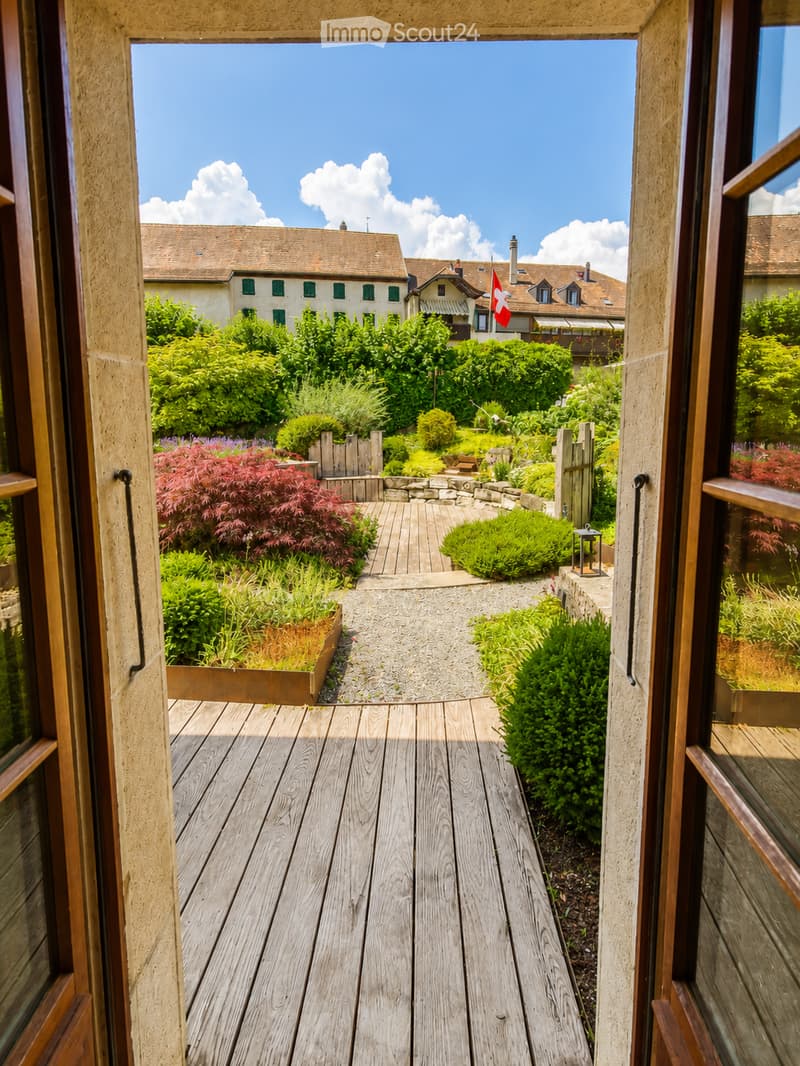A wooden deck leads to a well-maintained garden with various plants, shrubs, and a pathway, with a view of neighboring houses and a Swiss flag in the background.