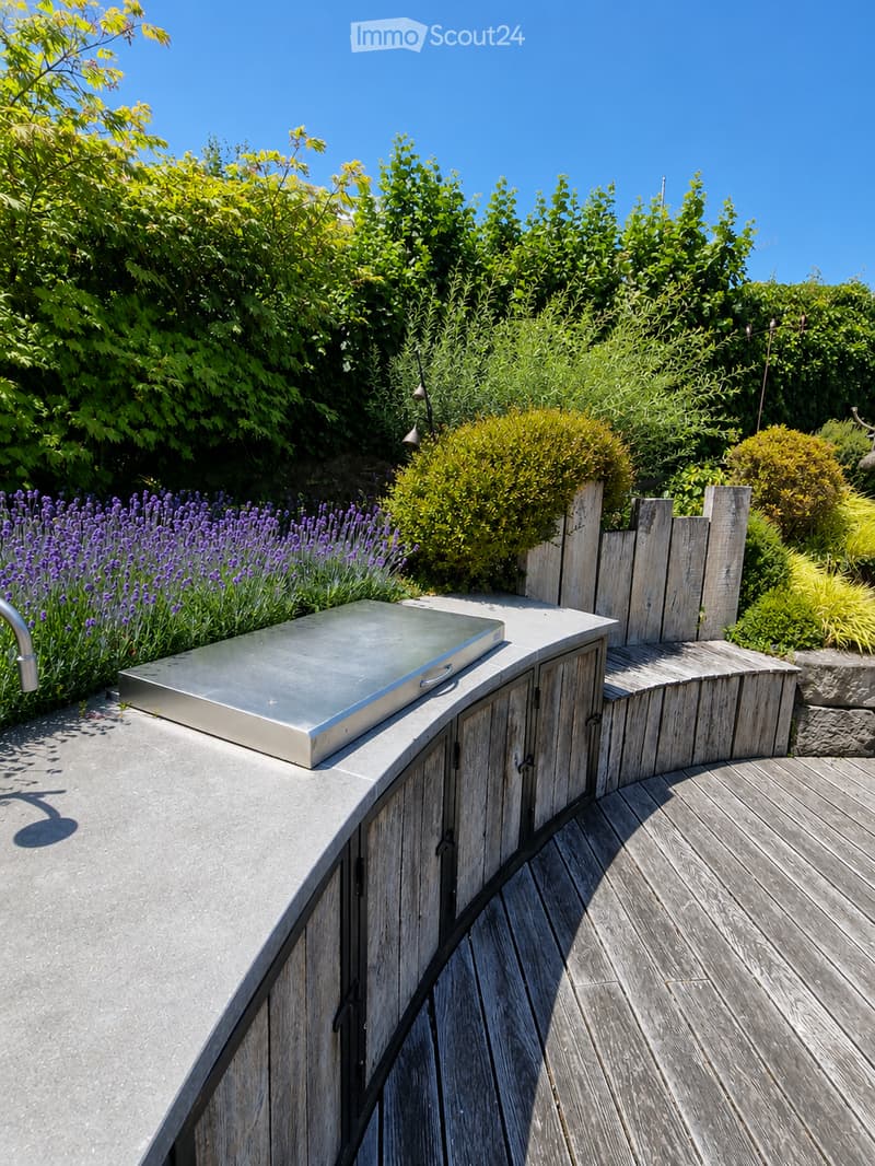 A modern outdoor kitchen area with a stainless steel grill, wooden cabinetry, and a wooden deck, surrounded by lush greenery and lavender plants.