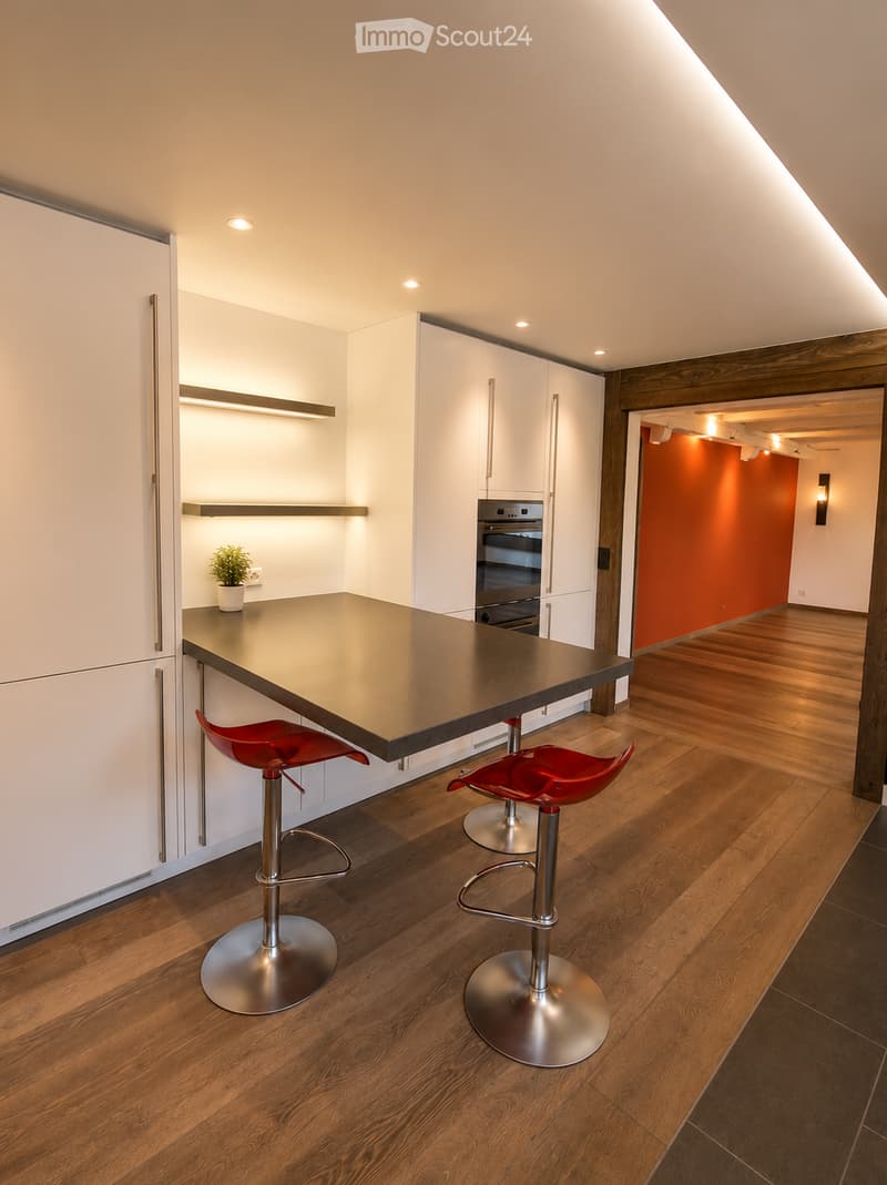 Modern kitchen with white cabinets, dark countertop, red stools, and built-in oven. Wooden flooring and recessed lighting.