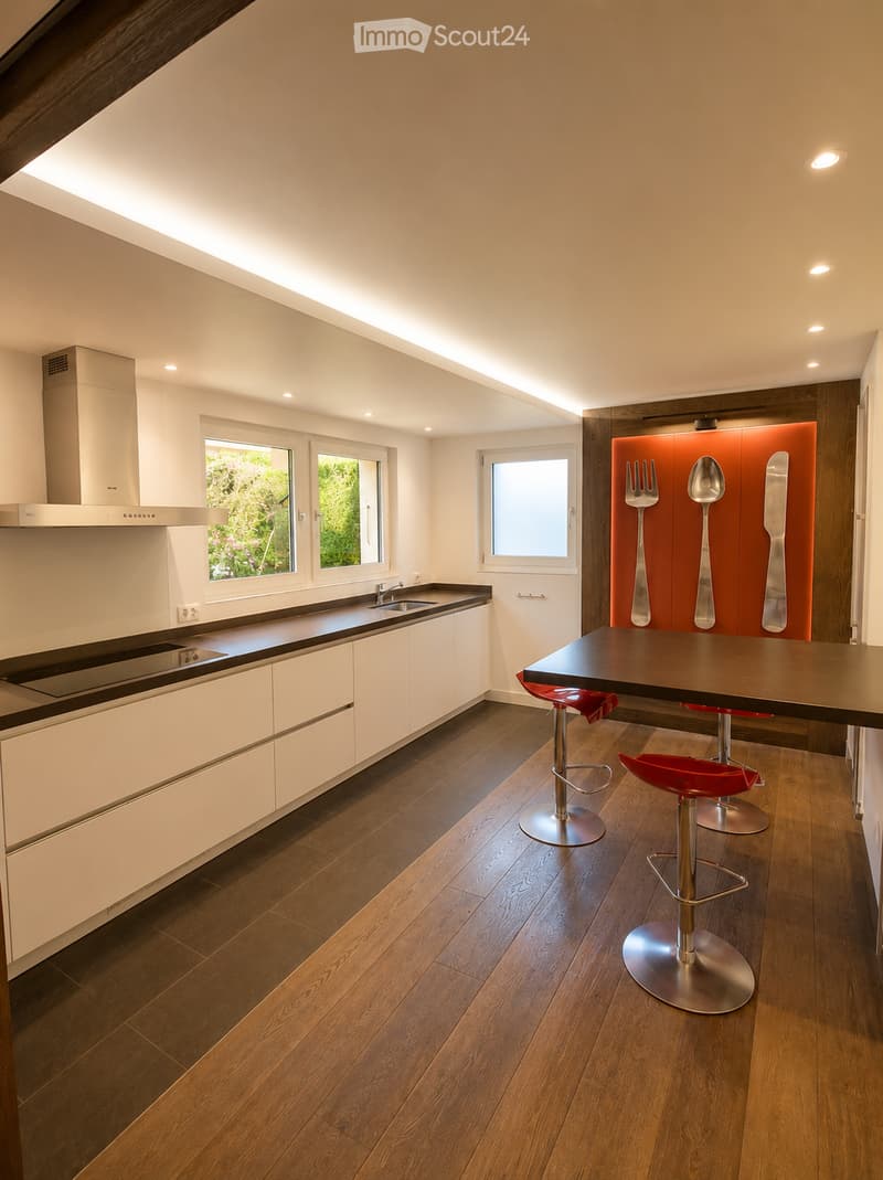 Modern kitchen with white cabinets, dark countertops, and a central island with red stools. Features include a range hood, sink, and large windows with a view of greenery.