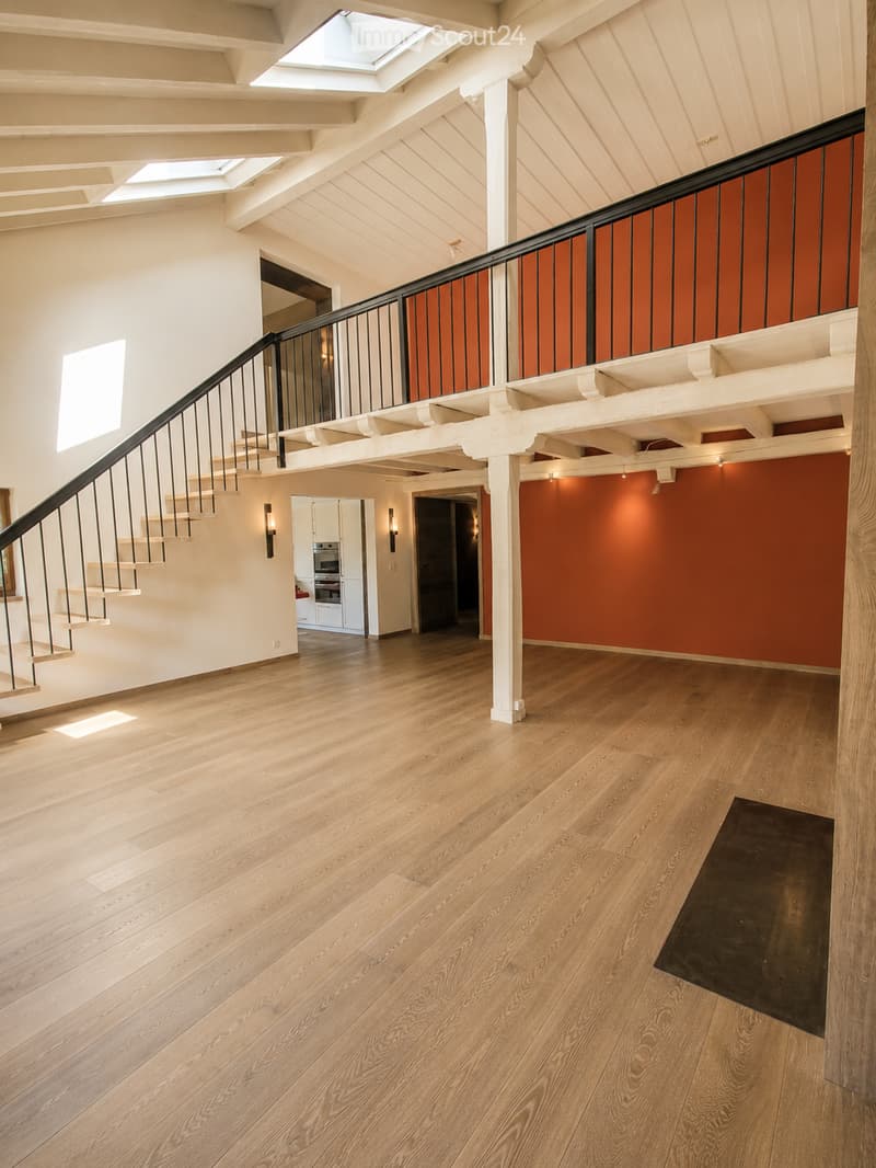 Spacious hallway with wooden flooring, white walls, and a staircase leading to an upper level. Features a black railing, warm lighting, and a glimpse of a kitchen area with white cabinets and stainless steel appliances.