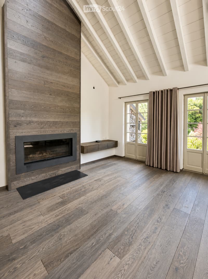 Modern living room with exposed wooden beams, a sleek fireplace, wooden flooring, and large windows with curtains.