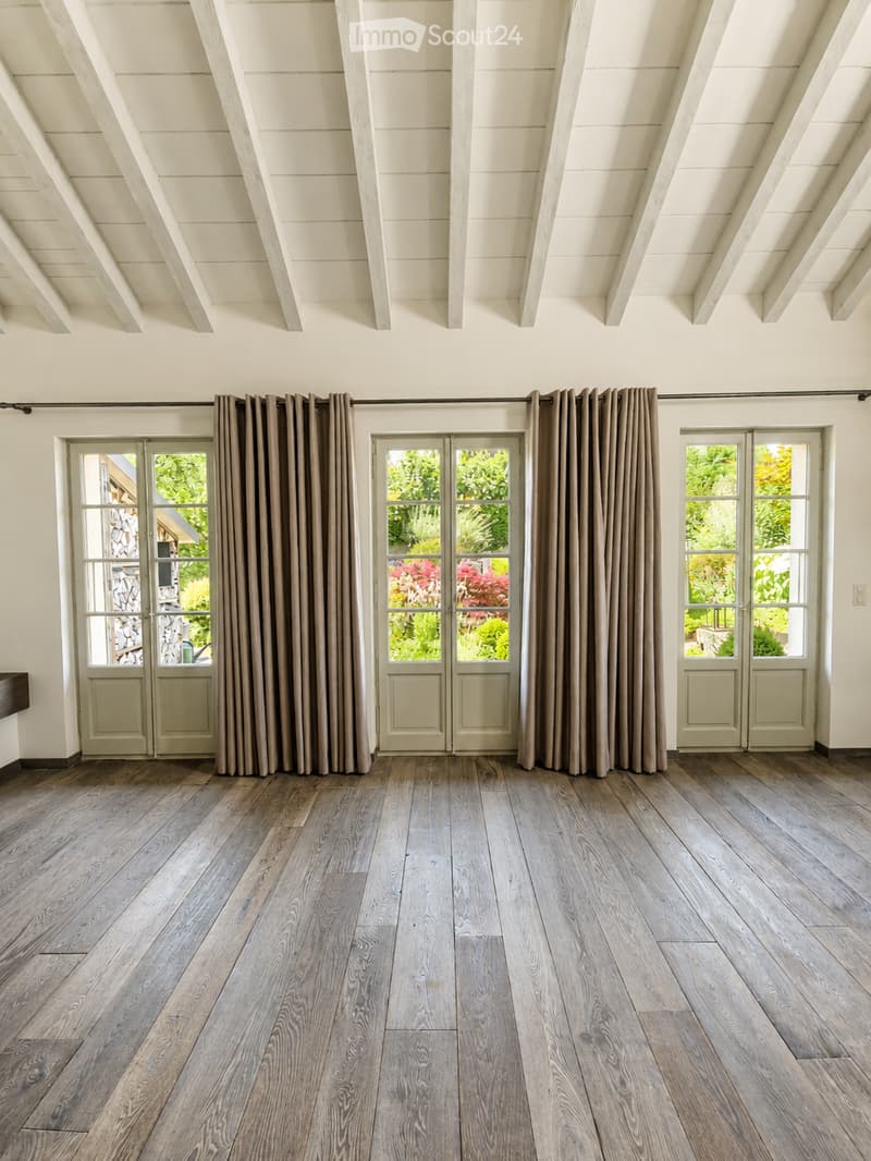 Empty room with wooden flooring, large windows with curtains, and a high, exposed wooden ceiling.