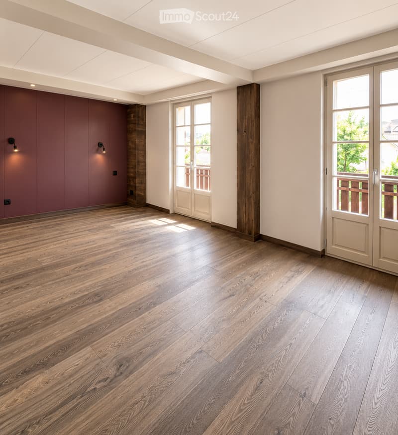 Empty room with wooden flooring, white walls, and large windows with white frames. The room features built-in wooden paneling on one wall and recessed lighting.