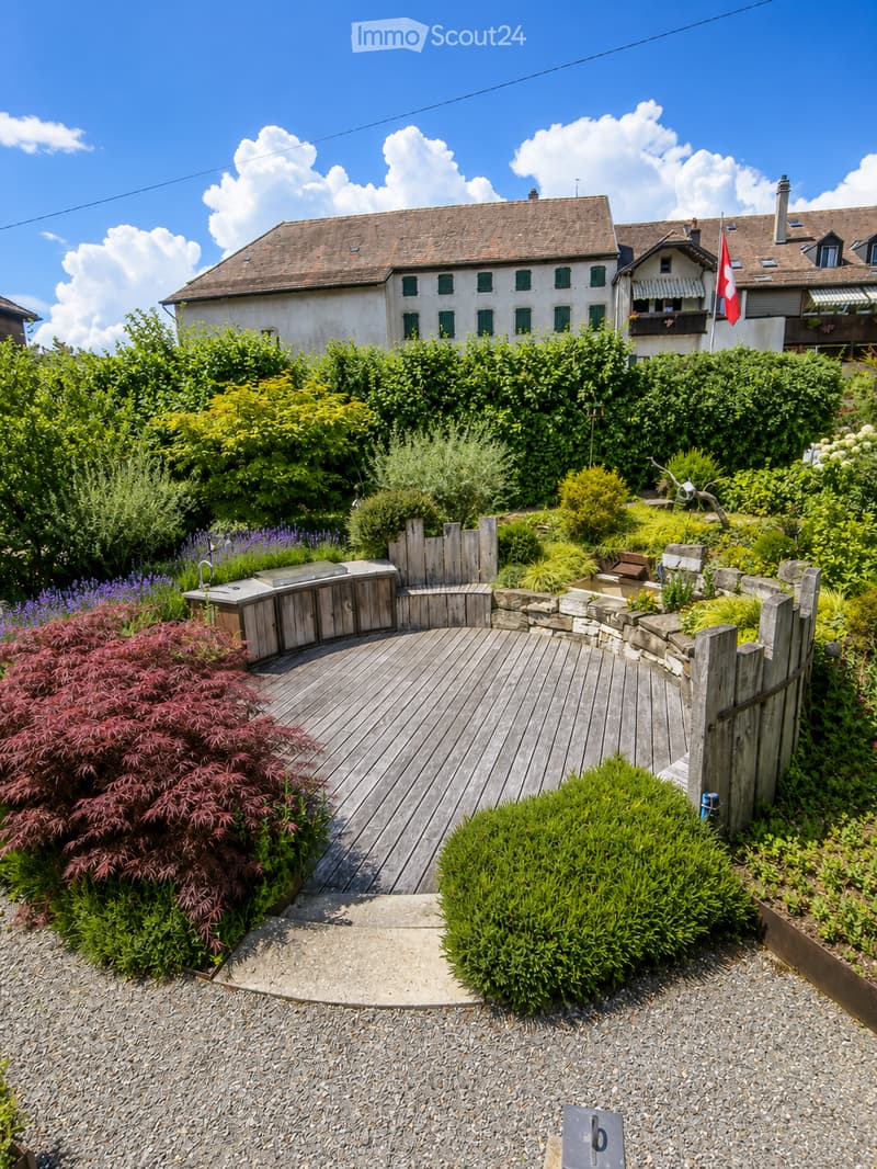 A well-maintained garden with a wooden deck, stone walls, various plants, and shrubs. A small outdoor kitchen area is visible, and a Swiss flag is displayed on a nearby building.