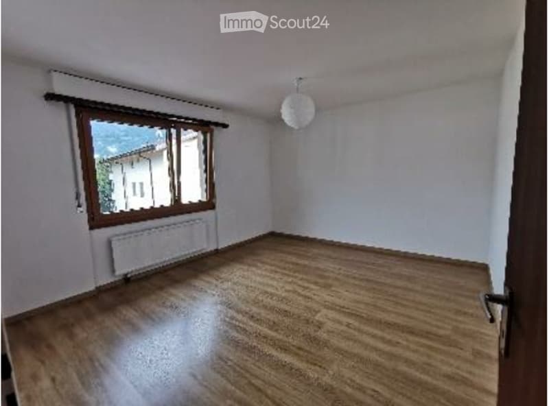 Empty room with wooden flooring, white walls, large window with wooden frame, radiator, and a hanging light fixture.