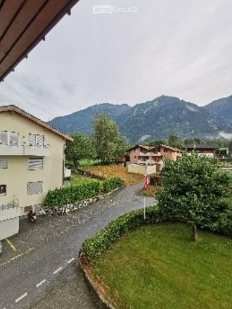 A view from a balcony or terrace overlooking a quiet residential area with houses, a driveway, and a mountain backdrop.