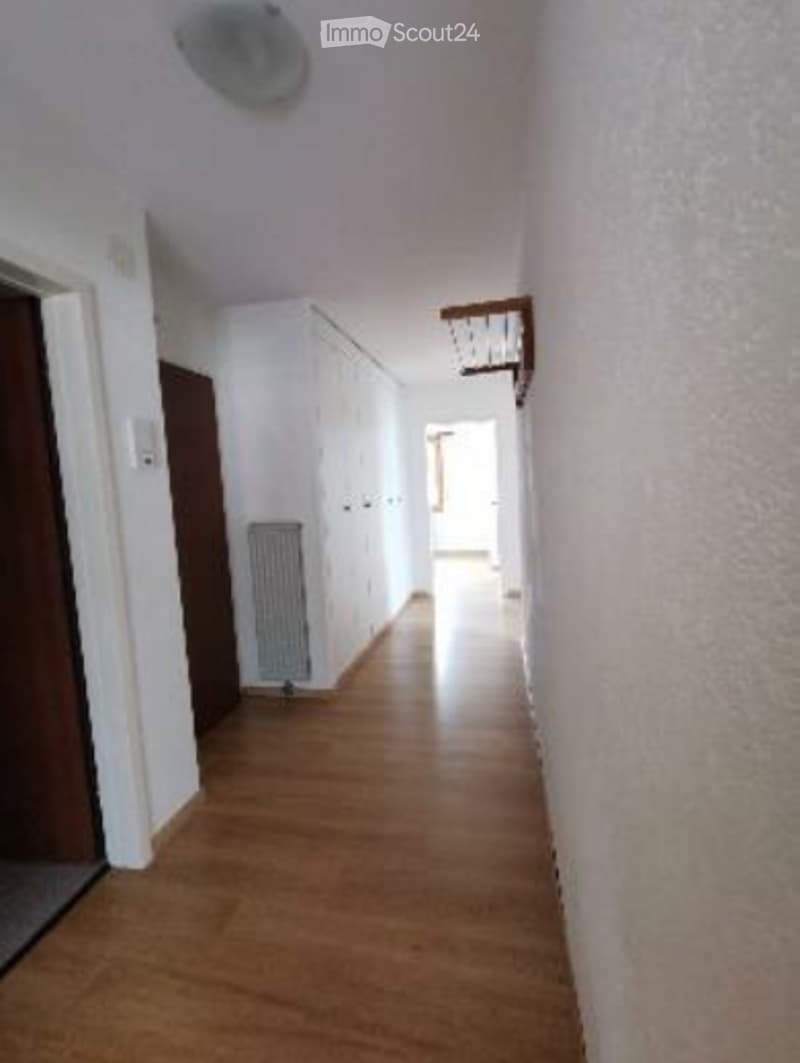 A long, narrow hallway with wooden flooring, white walls, and a radiator on the left side. A small shelf is mounted on the wall at the far end, and a ceiling light fixture is visible.