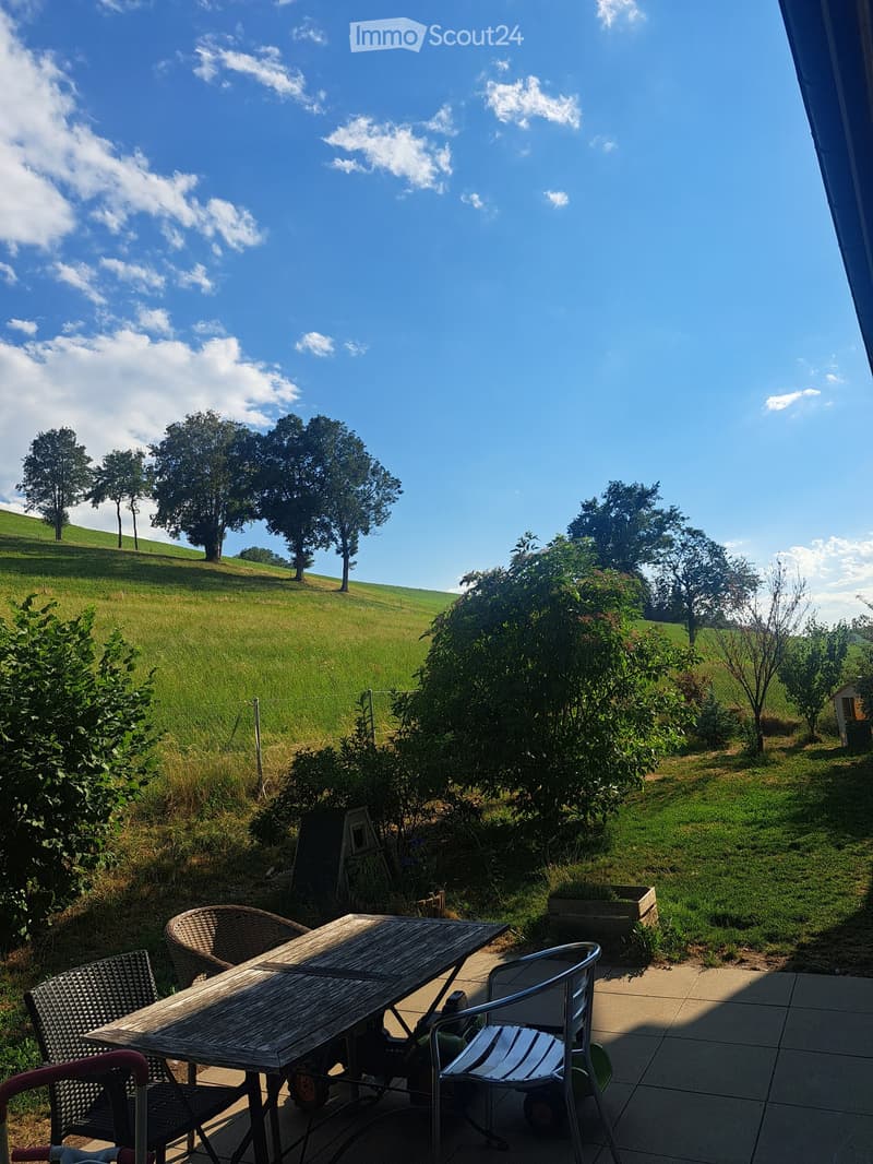 Outdoor patio with wooden table and chairs, surrounded by greenery and a scenic view of a grassy field and trees.