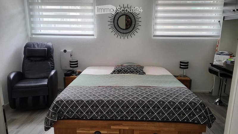 Cozy bedroom with a double bed, patterned duvet, nightstands with lamps, a black leather chair, and a decorative mirror on the wall.