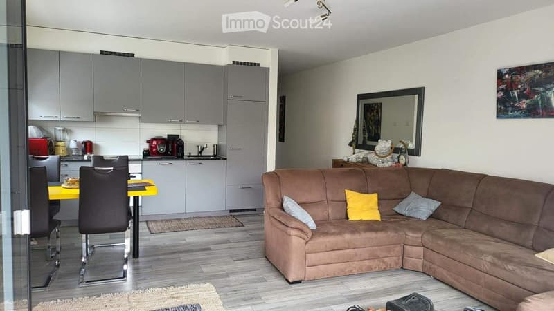 Modern open-plan living and dining area with a brown sectional sofa, yellow coffee table, dining table with chairs, and a small kitchen area with grey cabinets and various appliances.