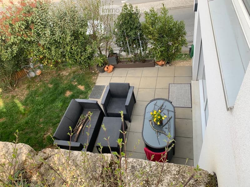 Balcony with a black wicker sofa set, a glass coffee table with a flower arrangement, and a small rug. The area is surrounded by greenery and has a stone wall on one side.