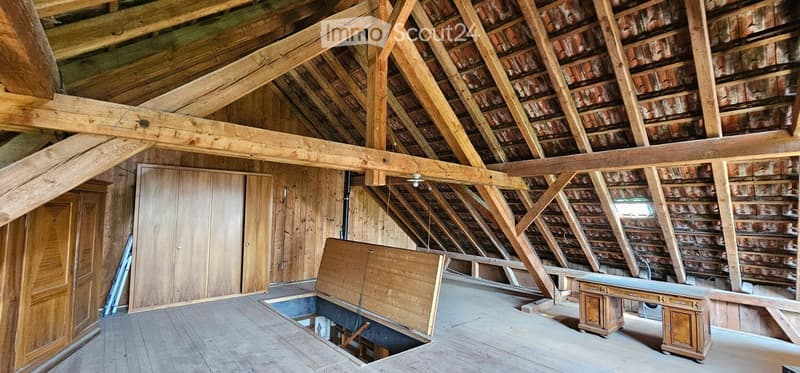 An attic space with exposed wooden beams and rafters, wooden walls, and a partially open floor revealing a drop-down staircase. The room is empty, with no furniture or furnishings.