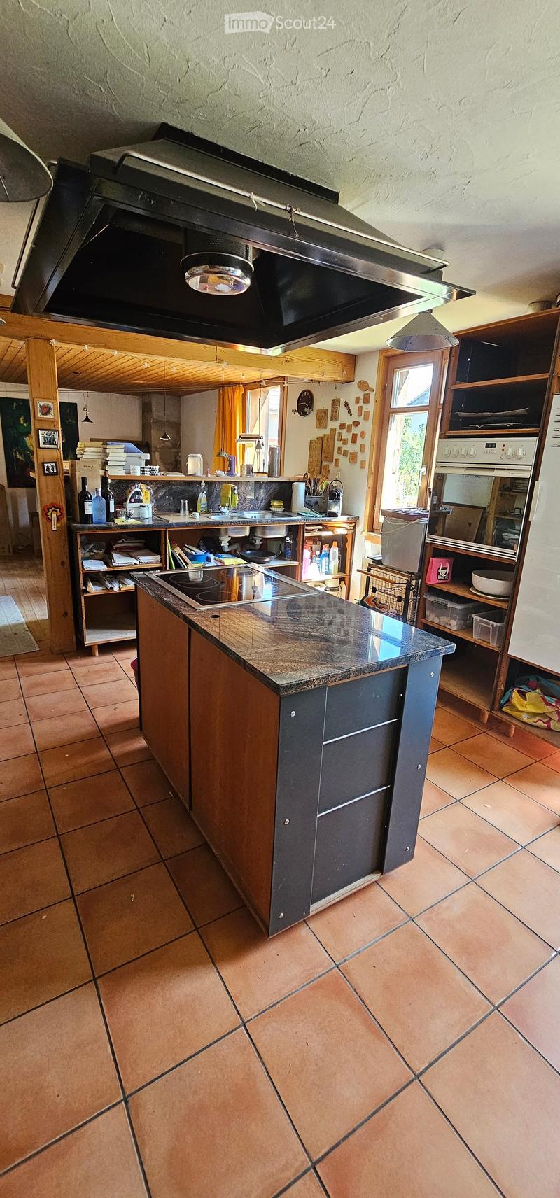 The kitchen features a central island with a granite countertop, a sink, and various appliances including an oven and refrigerator. Wooden cabinets and shelves are visible, along with various kitchen items and utensils.