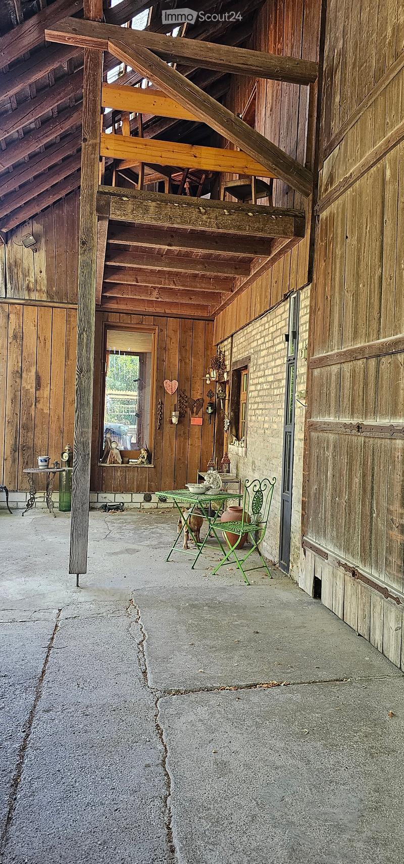 A rustic wooden corridor with exposed beams, wooden walls, and a concrete floor. Features include a green metal table and chair, a small table with a vase, and various decorative items on shelves.