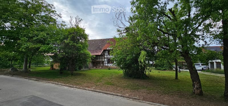 Traditional two-story house with a red-tiled roof, surrounded by lush greenery and trees. The house features a mix of wooden and plaster exterior walls, with a small porch area visible.