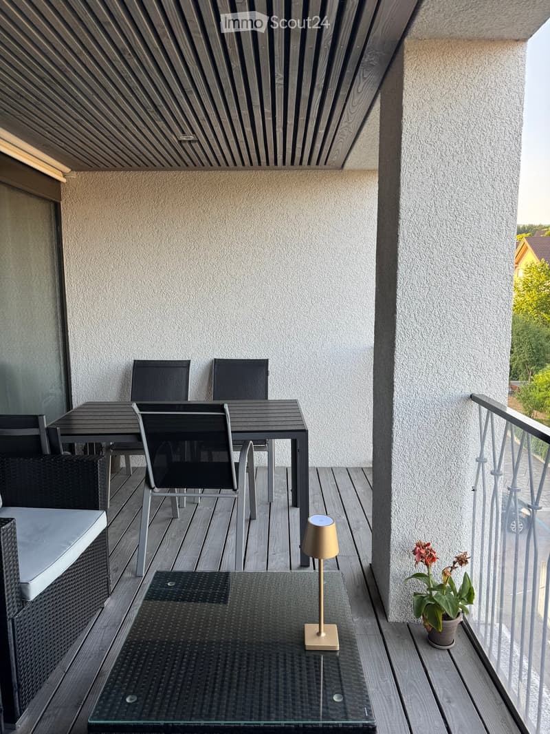 Balcony with wooden decking, glass table, chairs, lamp, and potted plant.