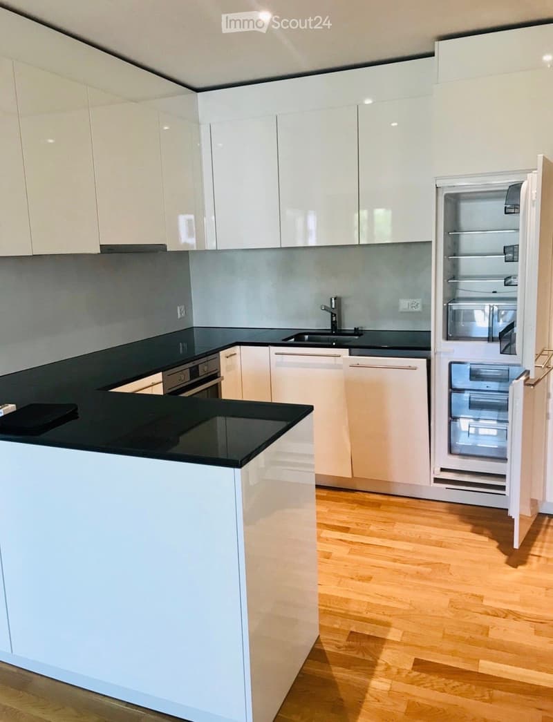 Modern kitchen with white cabinets, black countertops, and a central island. Features include a stainless steel sink, a built-in oven, and an open refrigerator.