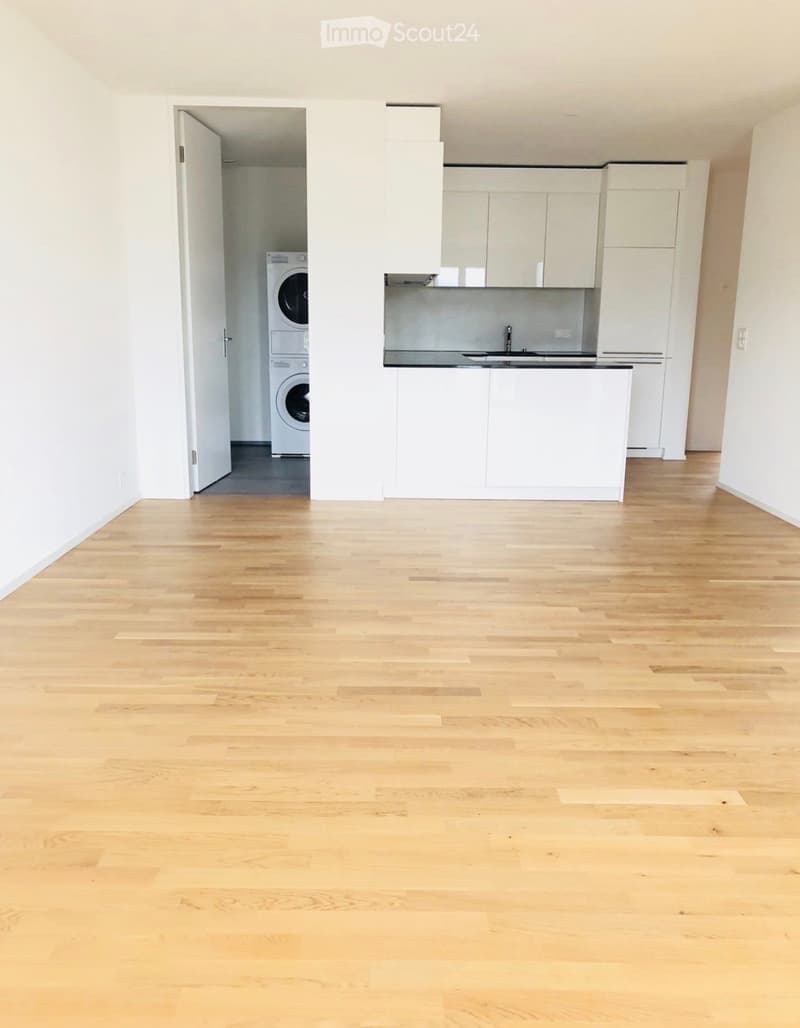 Empty room with light wooden flooring, white walls, and a partially visible kitchen area with cabinets and a washing machine in the background.
