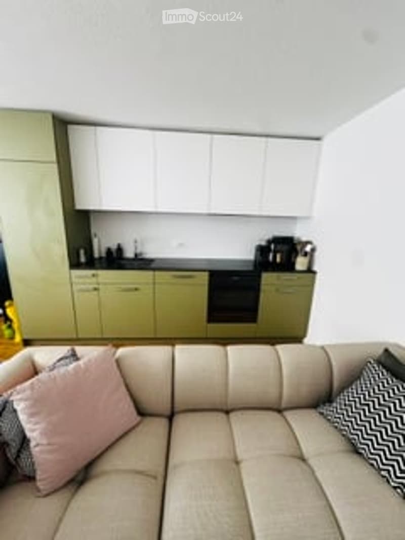 A modern, open-plan living and kitchen area. The living space features a light-colored sectional sofa with decorative pillows, while the kitchen area has sleek, light green cabinets and various appliances.