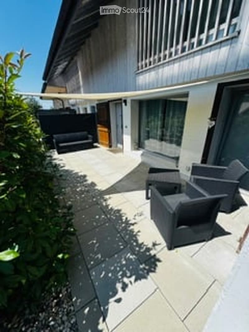 Modern outdoor patio with a stone floor, black lounge chairs, and a small black table. The area is surrounded by a low wall and has a view of a building with large windows and a balcony above.