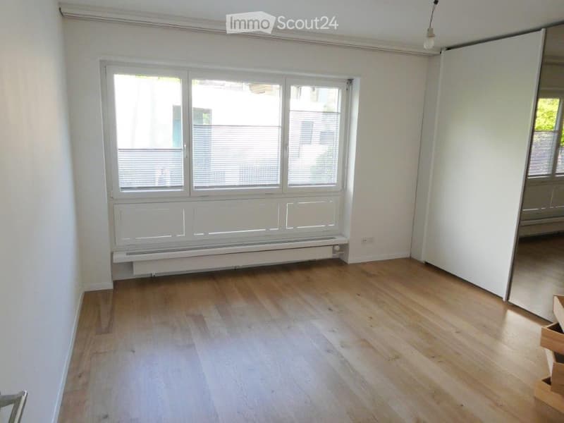 Empty room with wooden flooring, large window with blinds, radiator, and white walls.