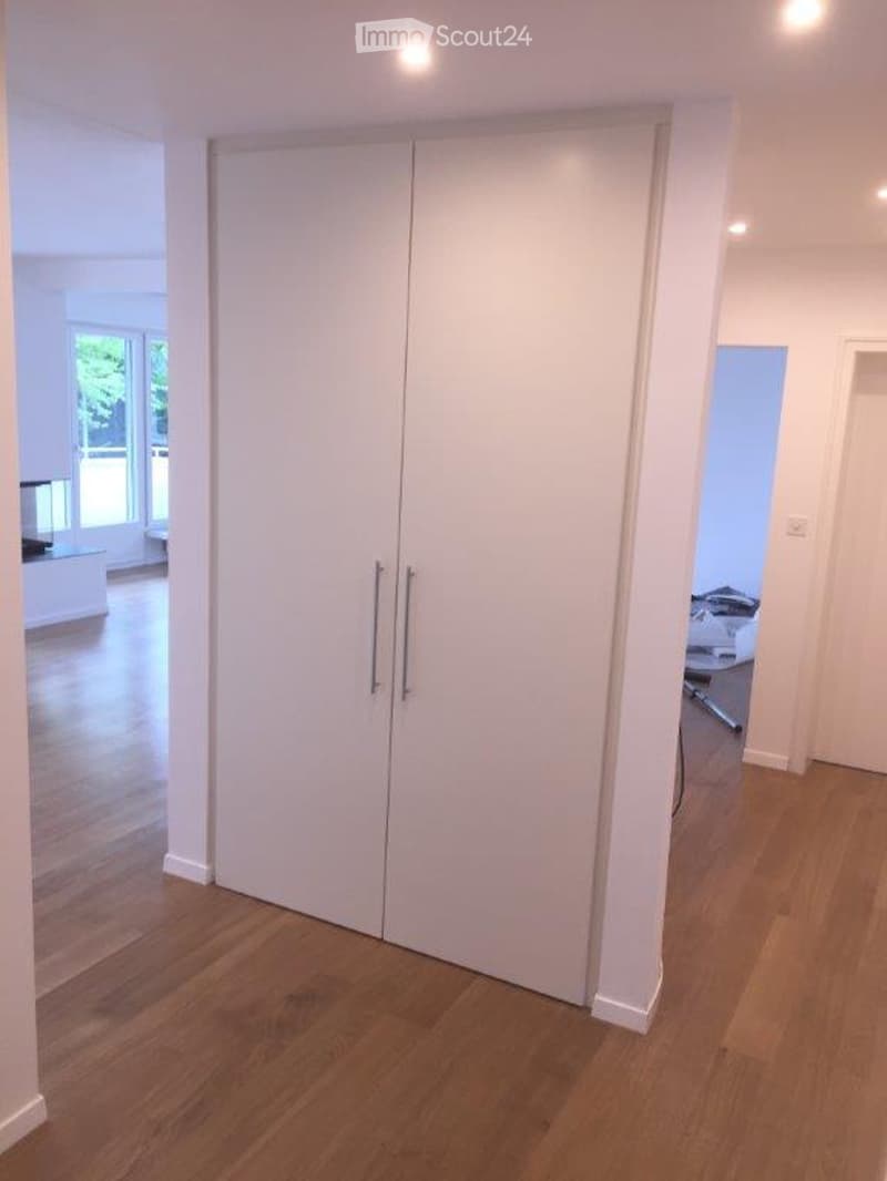 Empty room with wooden flooring and white walls, featuring a large white closet with double doors and silver handles. The room is well-lit with recessed lighting and has large windows allowing natural light to enter.