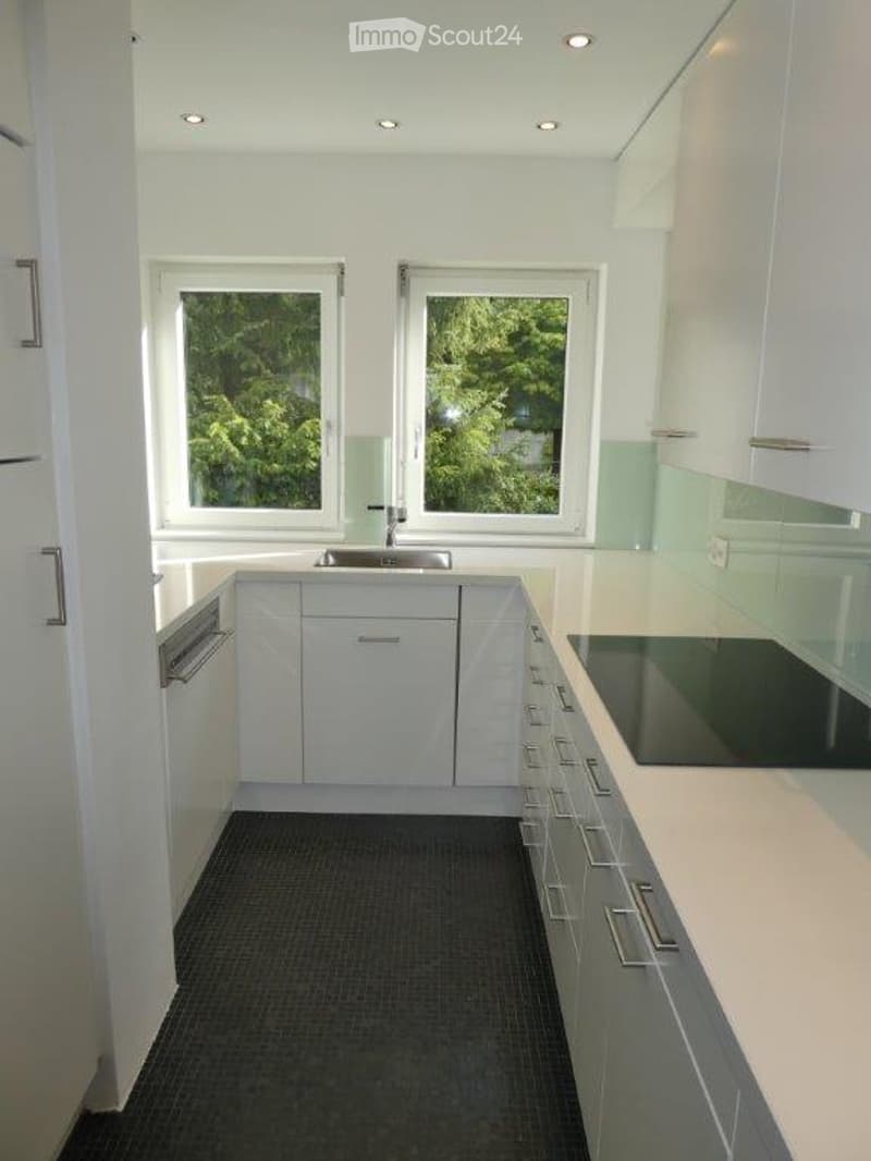 Modern kitchen with white cabinets, stainless steel appliances, and a dark tiled floor. Features a sink, dishwasher, and stove, with large windows offering a view of greenery.