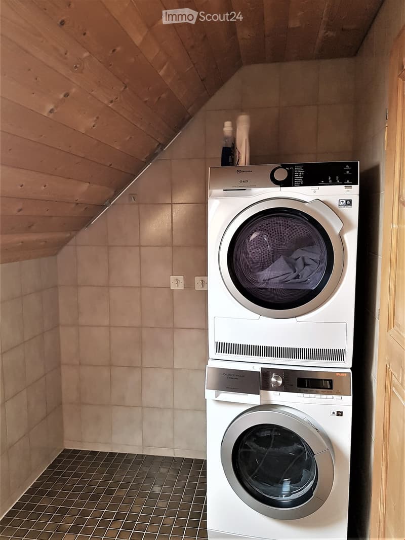 A small room with a washing machine and a dryer stacked on top of each other, tiled floor, tiled walls, wooden ceiling, power outlets