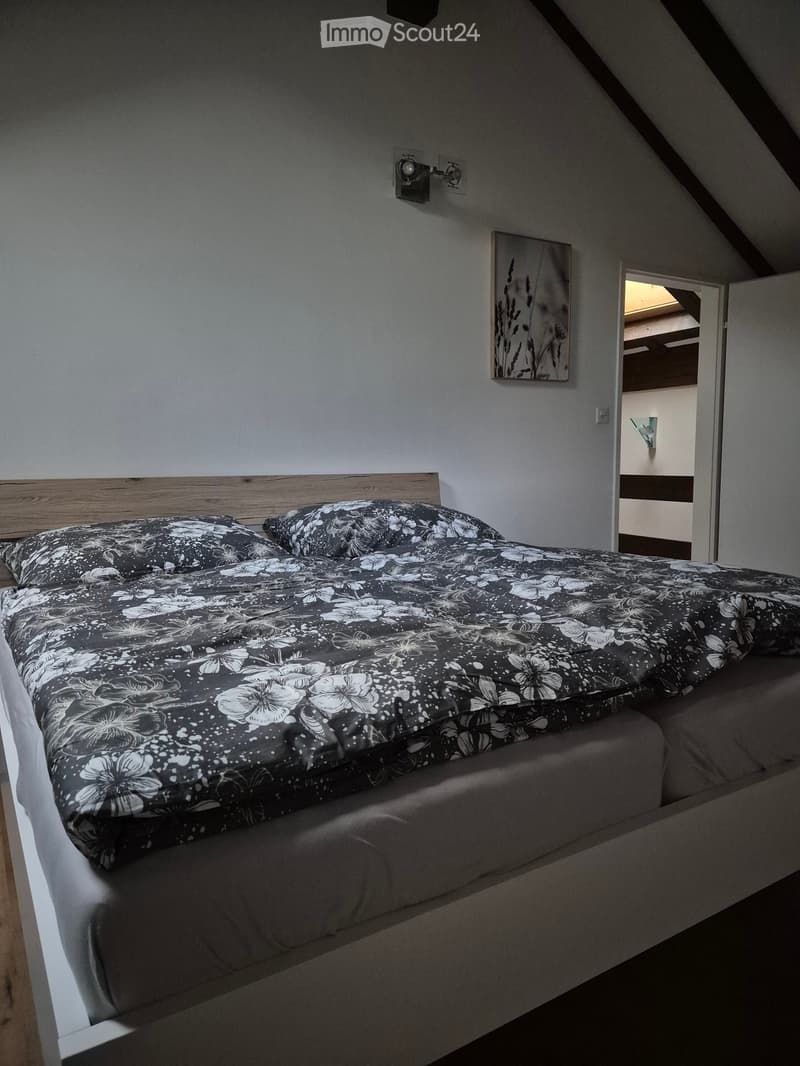 Bedroom with a double bed, floral-patterned duvet, wooden headboard, and a picture frame on the wall. The room has slanted ceilings typical of an attic space.