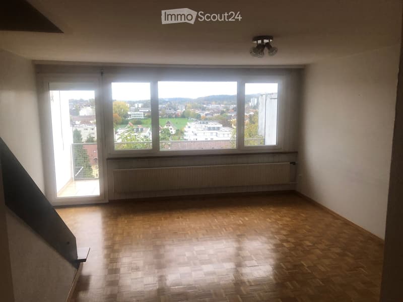 Empty room with wooden flooring, large windows offering a view of the city, radiator, and a sliding door leading to a balcony.