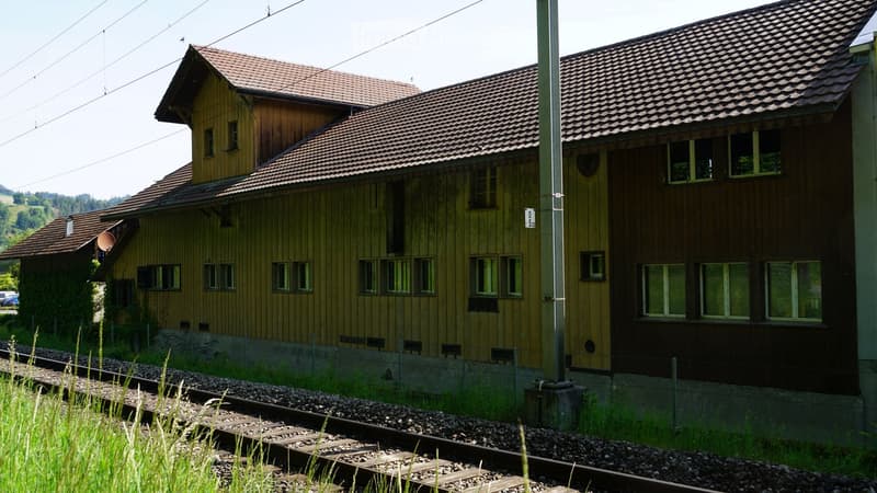 Two-story wooden building with a tiled roof, multiple windows, and a satellite dish on the left side. Located near train tracks with some overgrown vegetation in the foreground.