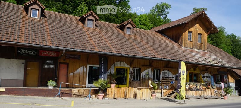 Two-story wooden building with a tiled roof, multiple windows, and signage for various businesses. The building has a covered entrance area with potted plants and banners.