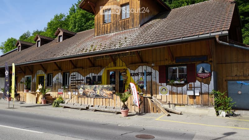 A rustic wooden building with a tiled roof, signage for "Kraftwerk" and "Coiffeur Mangstuli", outdoor seating area with wooden benches, potted plants, and banners.