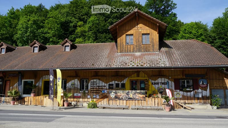 Two-story wooden building with a tiled roof, multiple windows, and a storefront with signage and banners. The building has a rustic design with wooden walls and a covered entrance area.