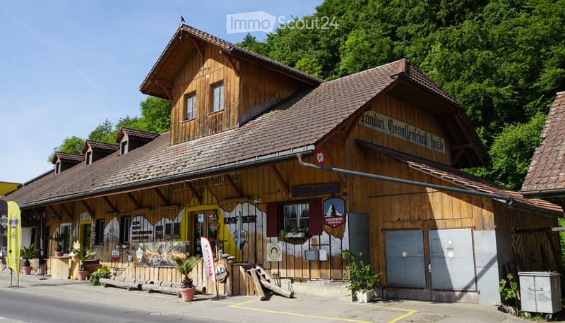 A rustic wooden building with a traditional design, featuring a sign that reads "Landau Gemüldenhausle" and a yellow entrance. The building has multiple windows, a covered porch area, and is surrounded by greenery and potted plants.