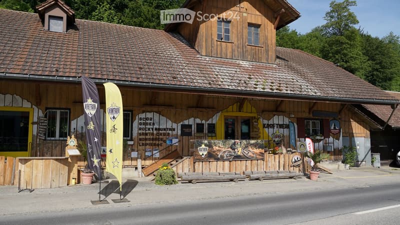 A rustic wooden building with a tiled roof, featuring outdoor seating, banners, and signage for a bike rental or cafe. The building has multiple windows and a covered entrance area.