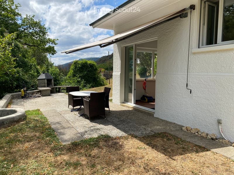 Outdoor patio area with a table and chairs, a covered awning, and a stone fireplace, surrounded by greenery and a scenic mountain view.