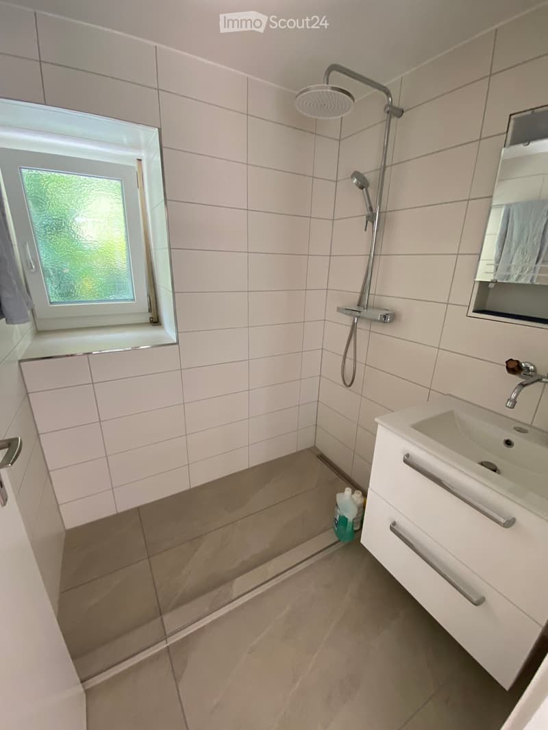 Modern bathroom with white tiles, a shower area, a sink with a mirror, and cleaning supplies on the floor.