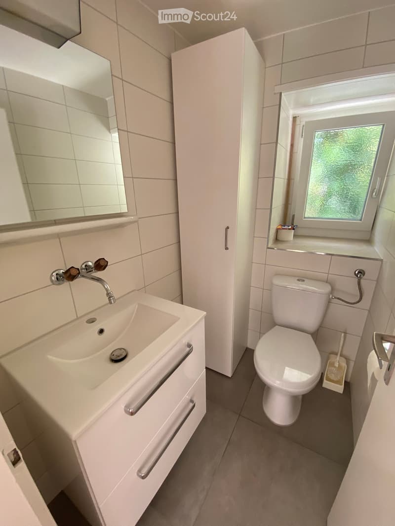 Modern bathroom with white tiles, a sink with a chrome faucet, a toilet, a mirrored cabinet, and a window with frosted glass.