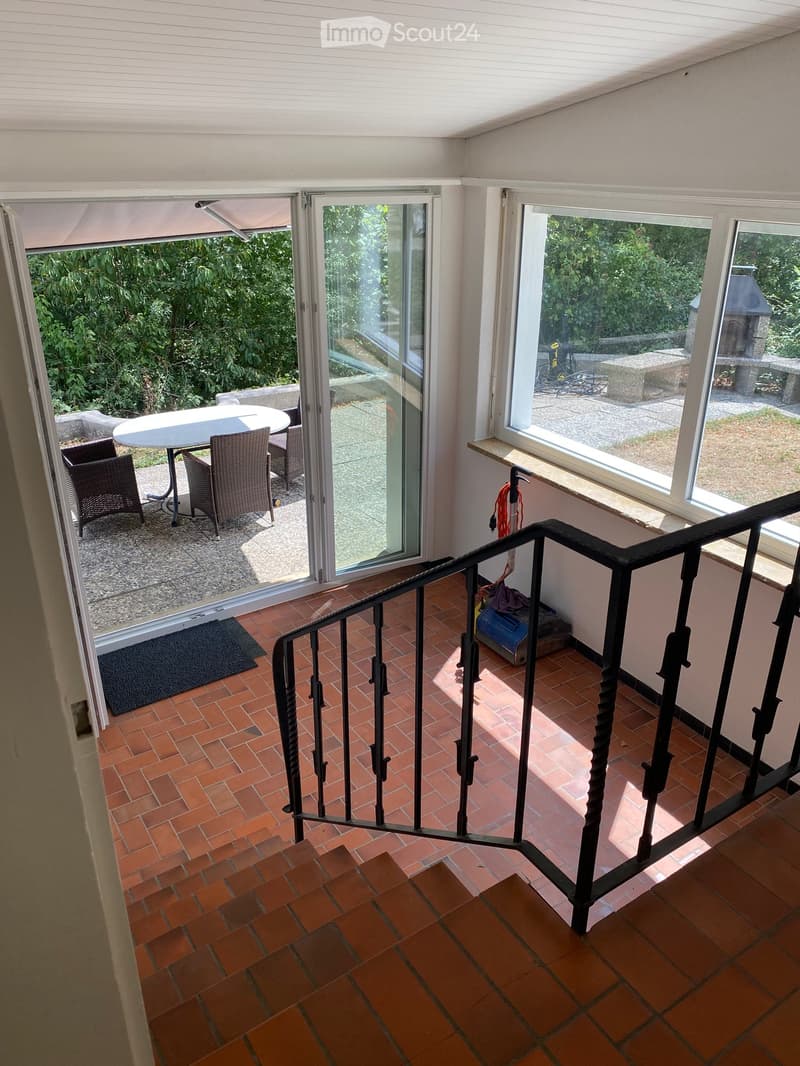 Balcony with brick tiles, black metal railing, and a small seating area with a table and chairs, overlooking a garden.