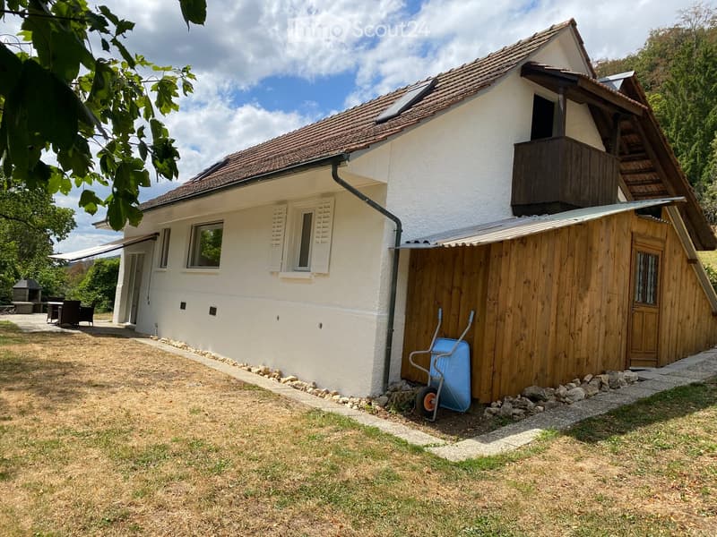 Two-story house with white exterior, brown tiled roof, and wooden balcony. Features include a covered entrance, a small garden area, and a wooden shed with a wheelbarrow.