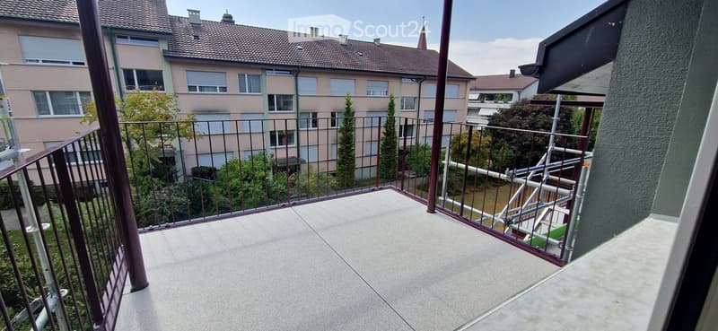 Balcony with metal railing, concrete floor, overlooking a residential area with buildings and trees.