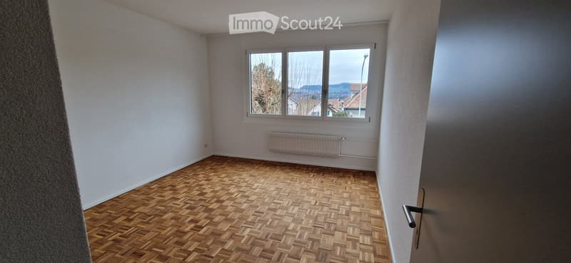 Empty room with wooden parquet flooring, white walls, and large windows offering a view of the outside. No furniture or equipment is present.