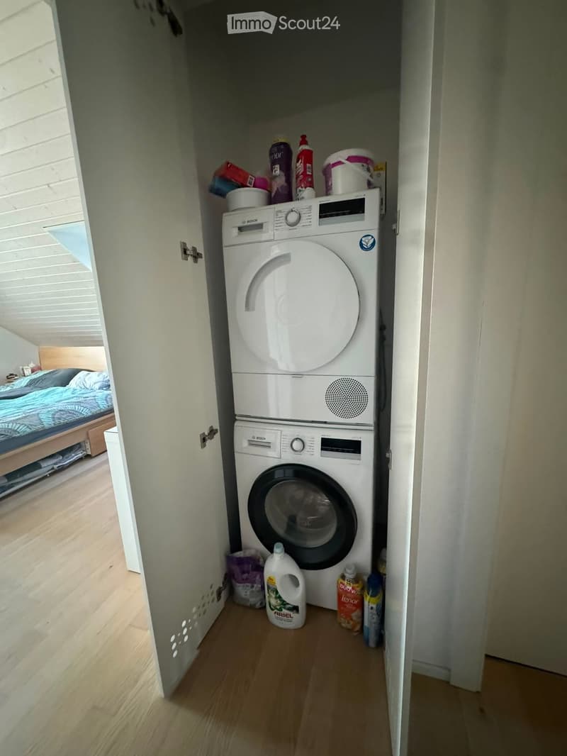 A small, compact laundry area with a stacked washing machine and dishwasher, surrounded by cleaning supplies and a glimpse of a bedroom with a bed and blue bedding.