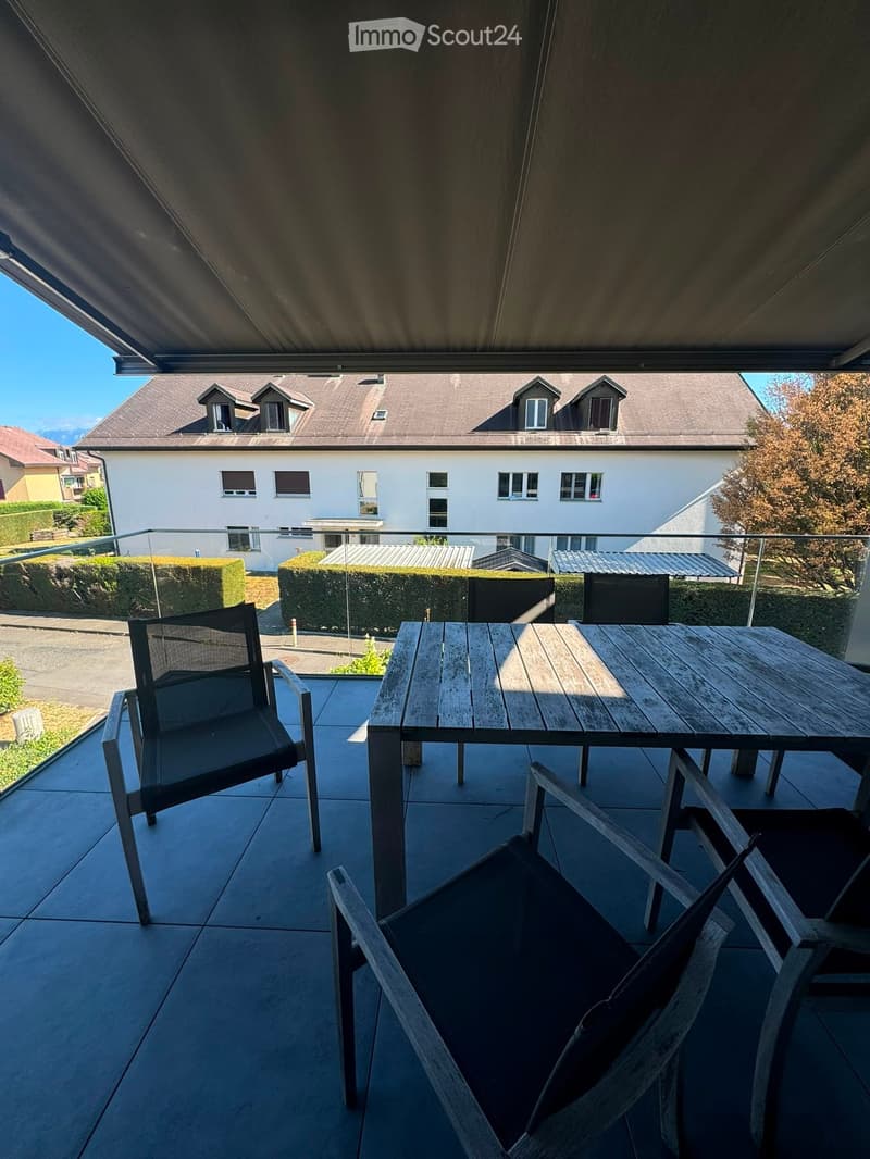 Balcony with a wooden table and two chairs, overlooking a residential area with houses and greenery.