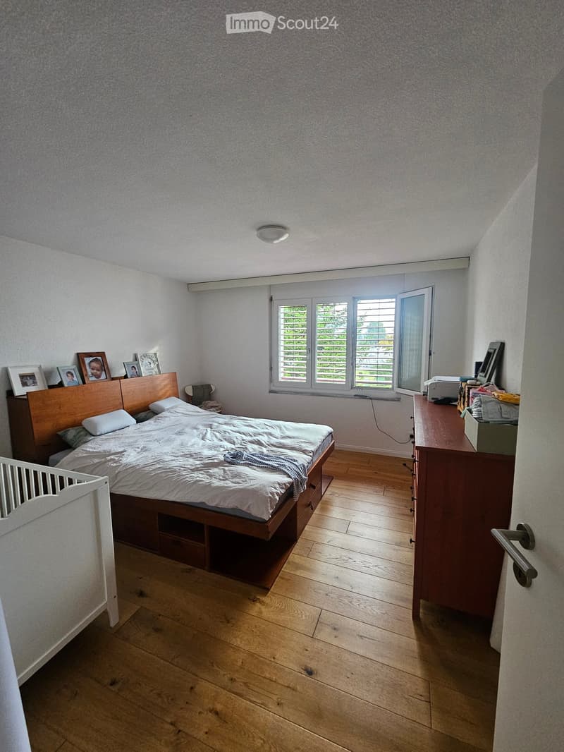 Bedroom with wooden floor, large window with blinds, bed with white bedding, nightstand with mirror, chair, baby crib, and picture frames on the wall.