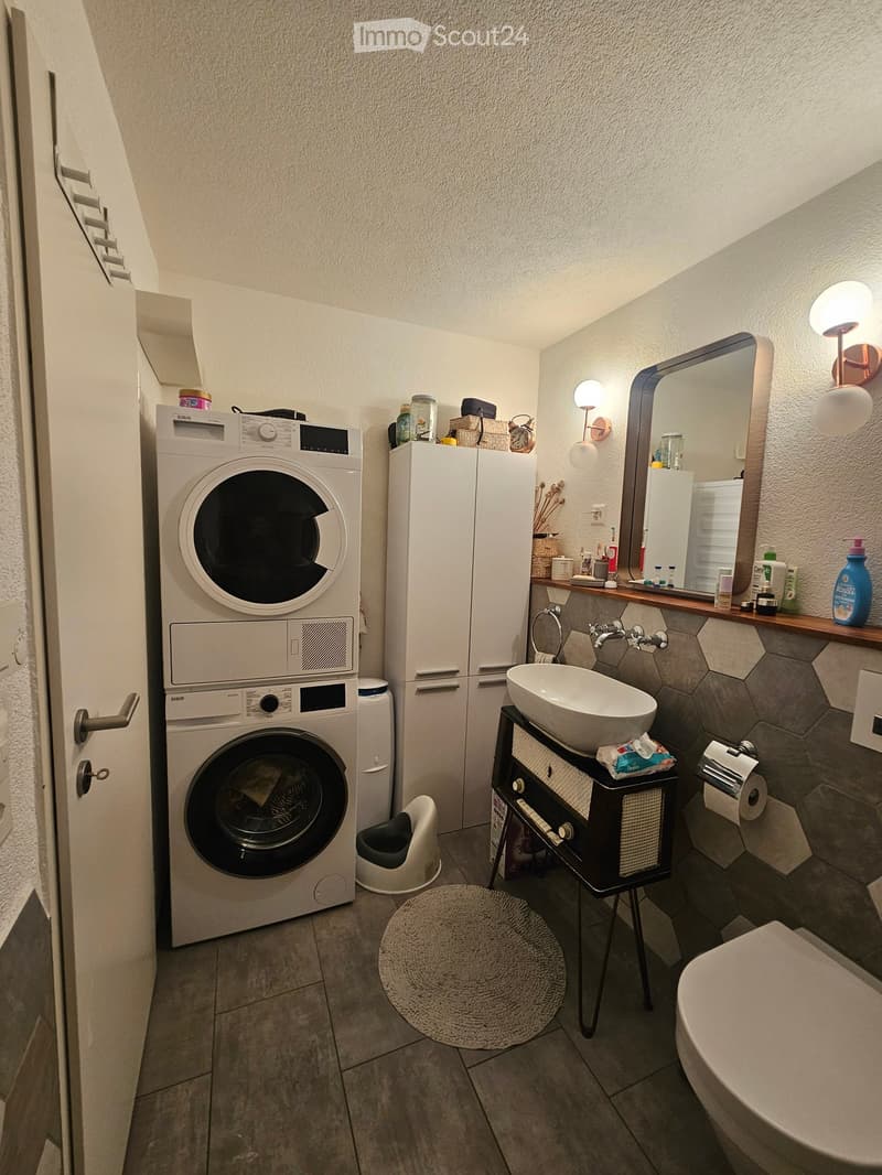 Modern bathroom with a washing machine, sink, mirror, and toilet. Features include a hexagonal tile backsplash, a round rug, and a small stool.