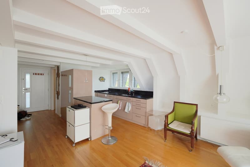 Bright, open-plan room with wooden flooring, a kitchen area with cabinets and a sink, a small bar stool, a green armchair, and a modern light fixture.