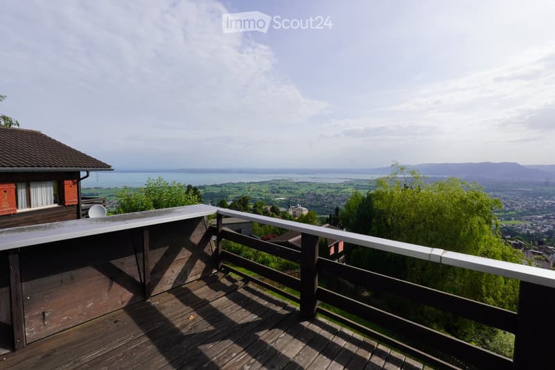 Wooden balcony with white railing, overlooking a scenic lake and distant town with surrounding greenery.